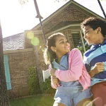 Two young kids on a swing in front of a house