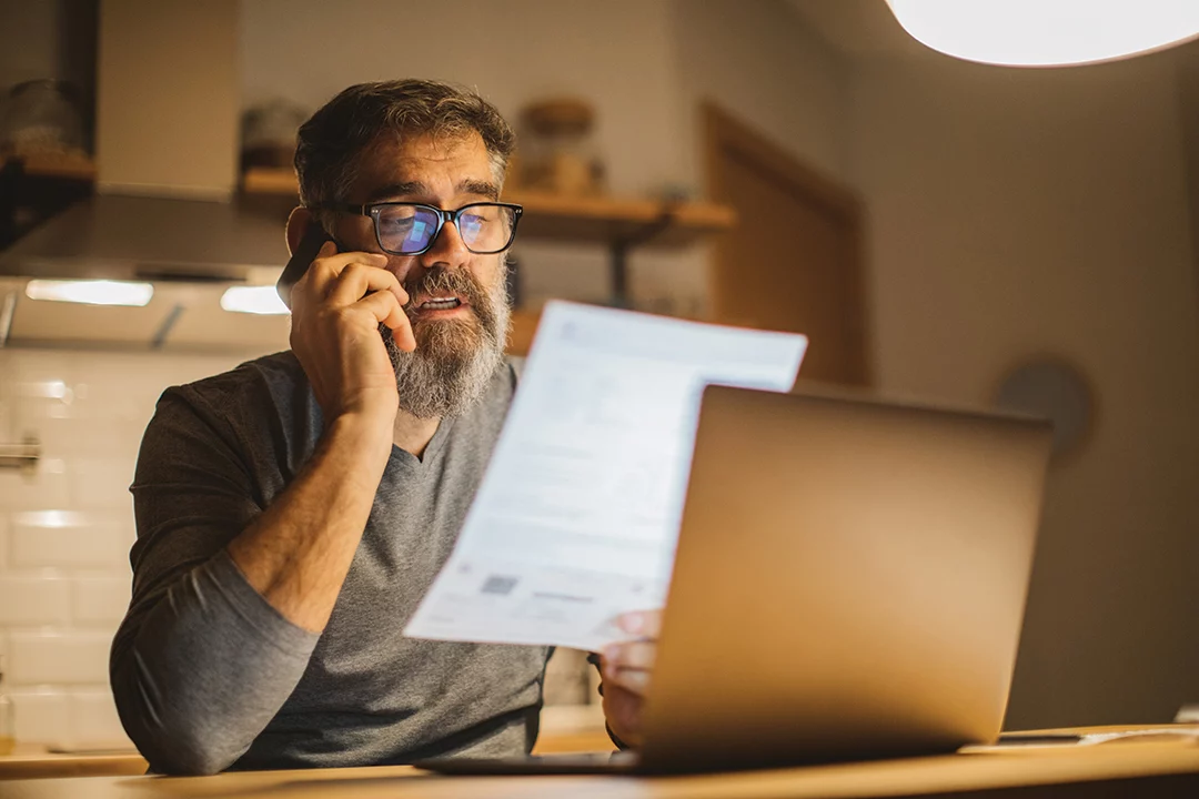 A man on the phone while looking a piece of paper with a laptop