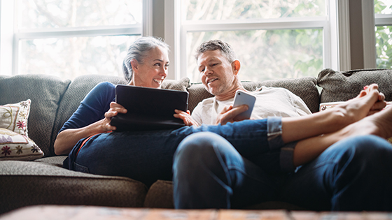 Couple relaxing on couch with one holding a tablet and one holding a smartphone.