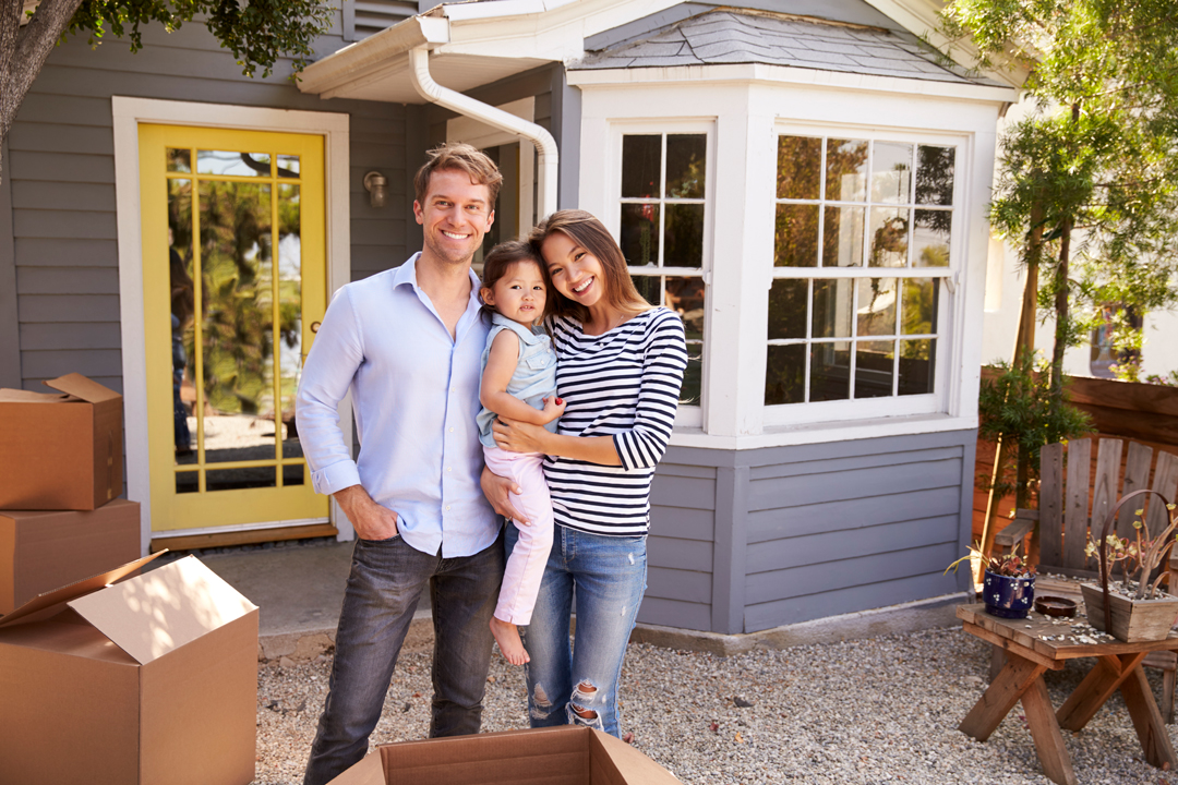 Smiling, young man and woman holding their daughter while standing in front of a new home surrounded by moving boxes.