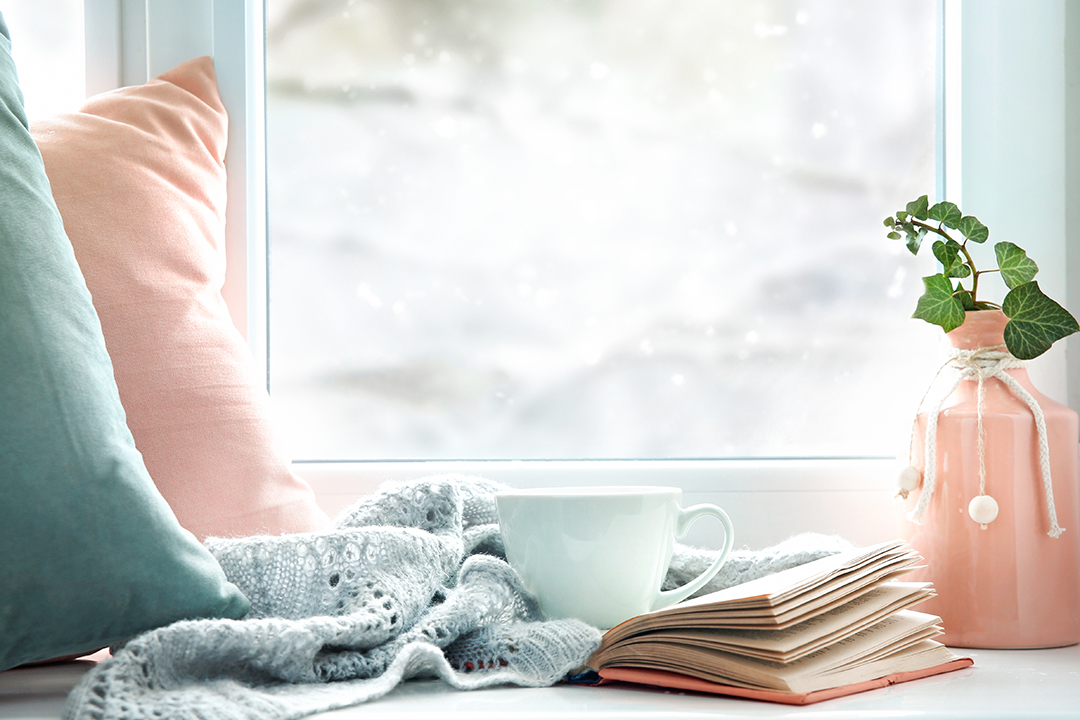 A mug, blanket,book, and pillows in front of a window with a snowy background